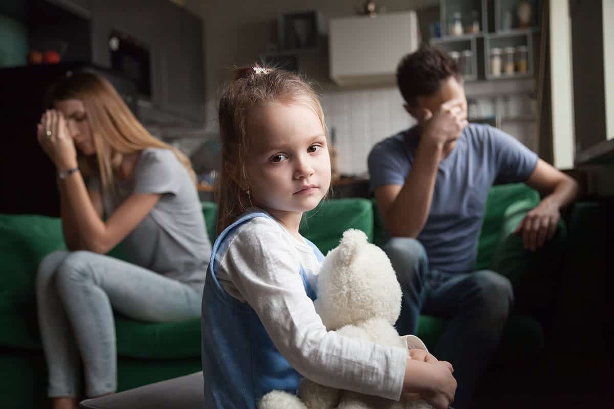 two parents in the background cover their faces while a child looks at the camera somberly as she experiences how Drugs affect family